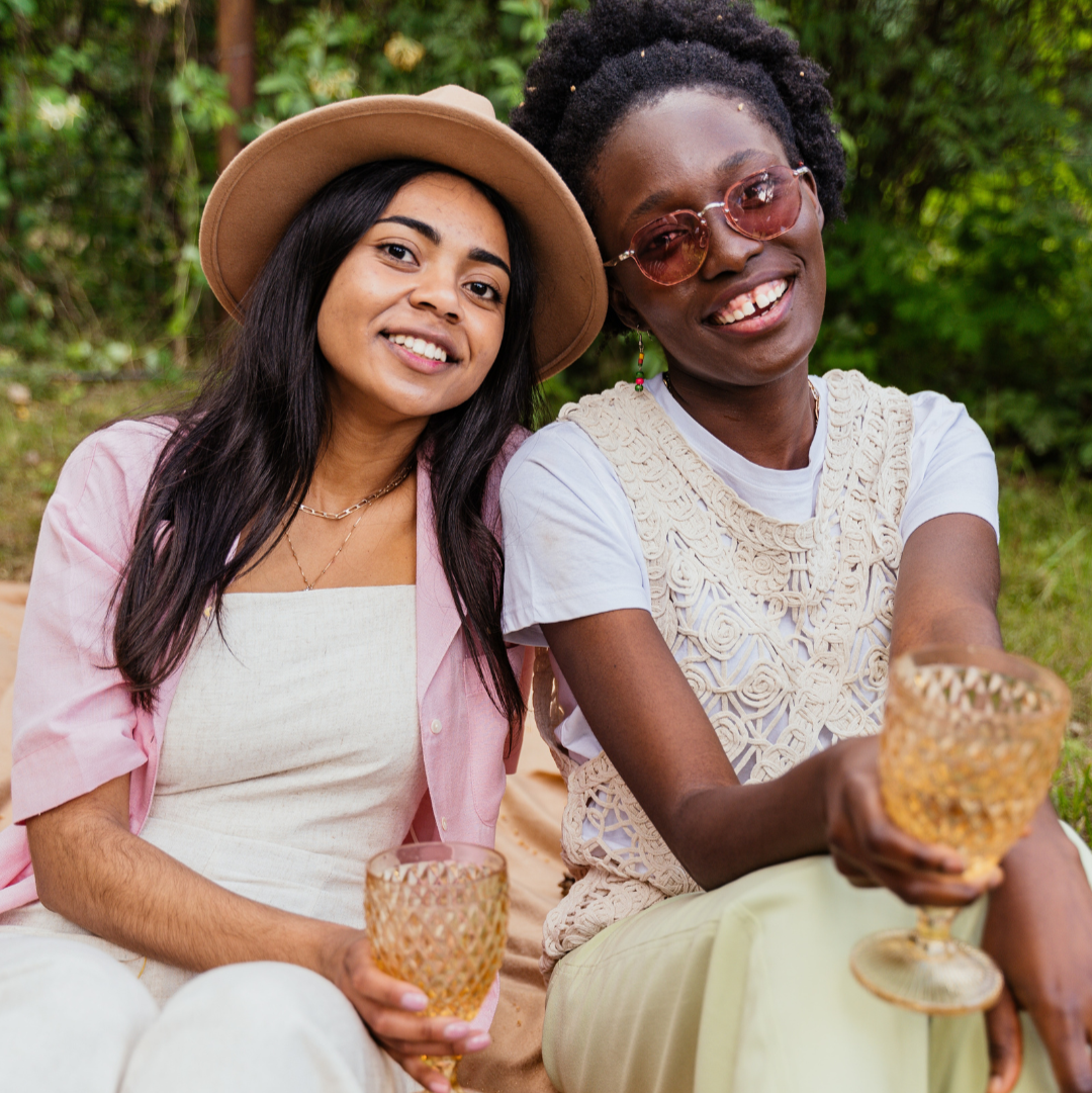 Two women sitting outside in the sunlight, smiling and clinking glasses of Nouvie sparkling wine, enjoying a joyful moment together.