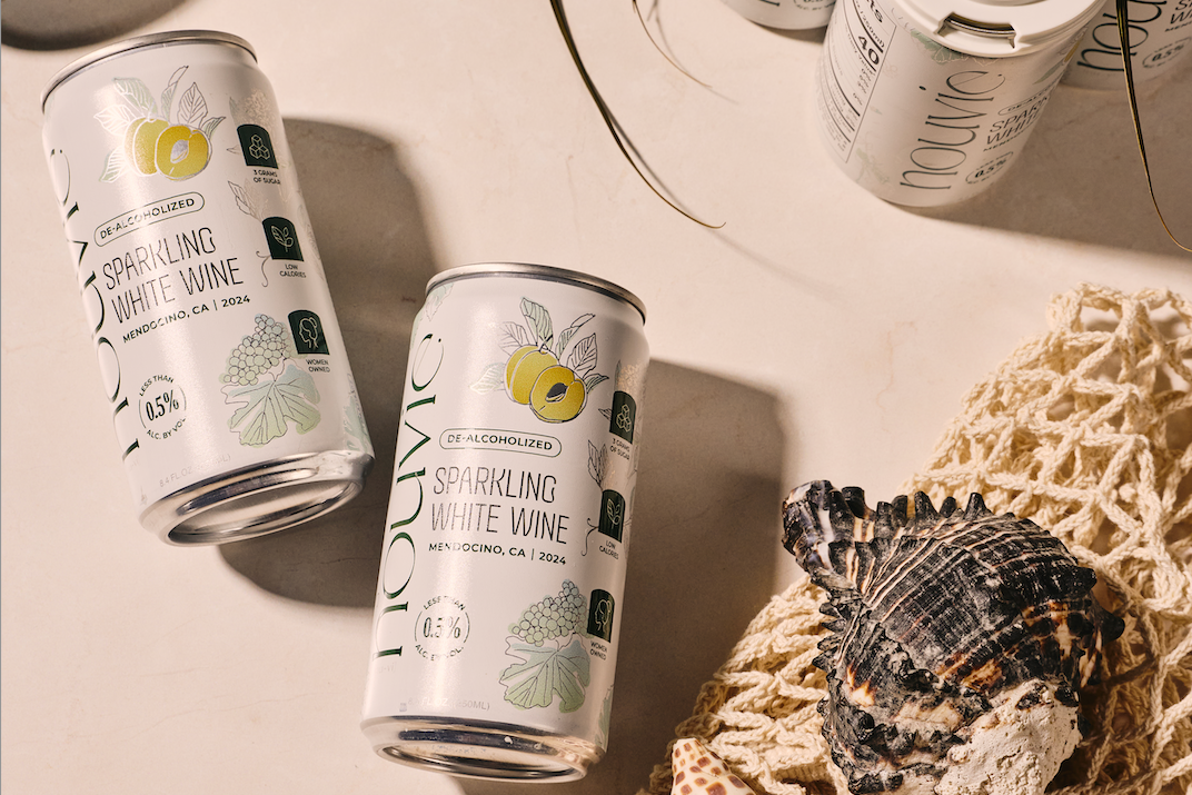 Two cans of KAVOS Sparkling White Wine are arranged on a light beige surface with beach-style props including black sunglasses, a white six-pack of KAVOS cans, seashells on a cream net, and green leaves, under warm directional sunlight.
