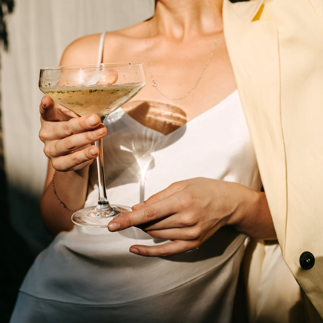 Woman in a white dress and yellow jacket holding a glass of De-Alc Nouvie Spanish sparkling white wine, standing in the sunlight 